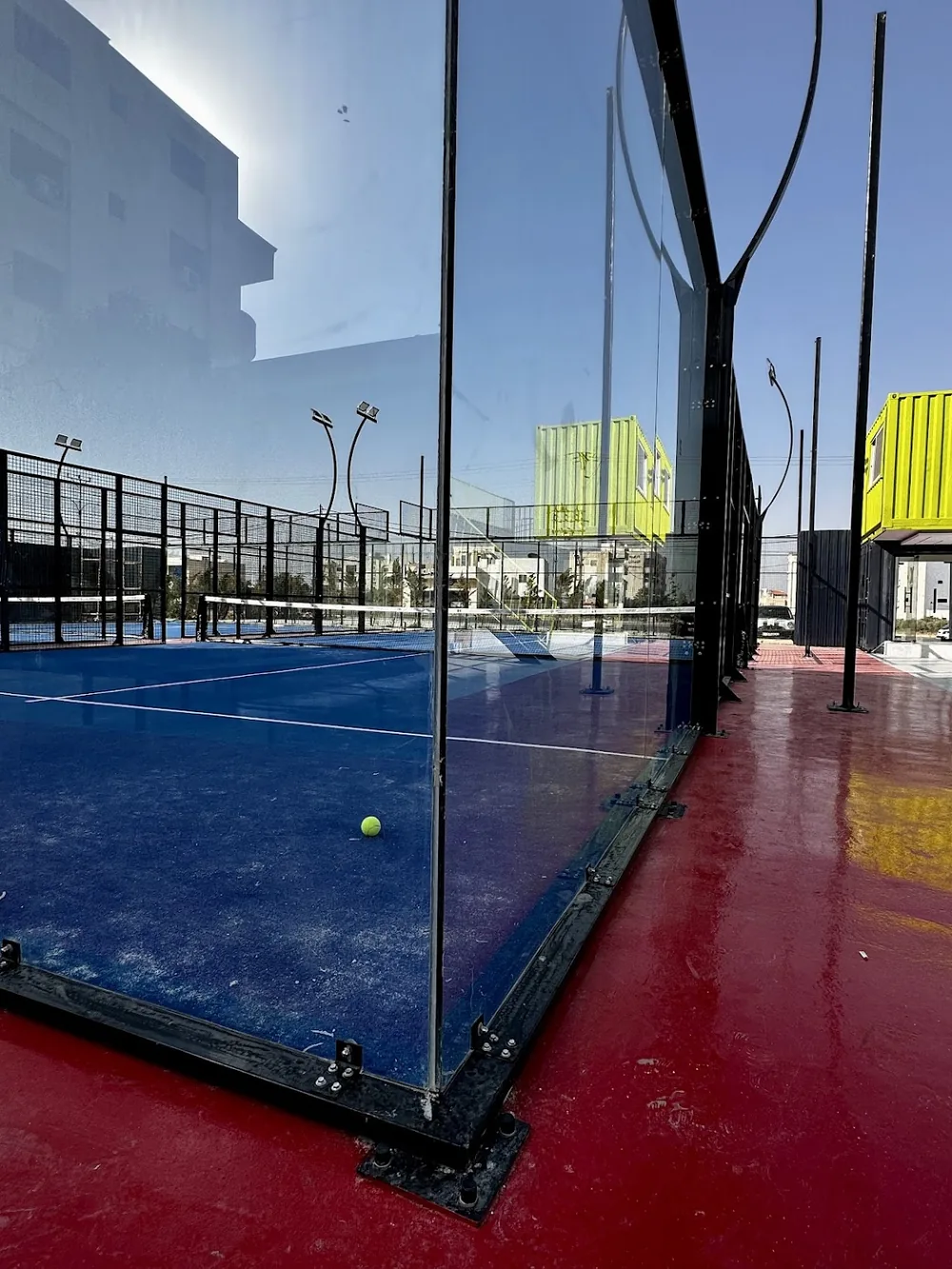 A ball resting on the blue turf beside the glass wall of a Padel X court